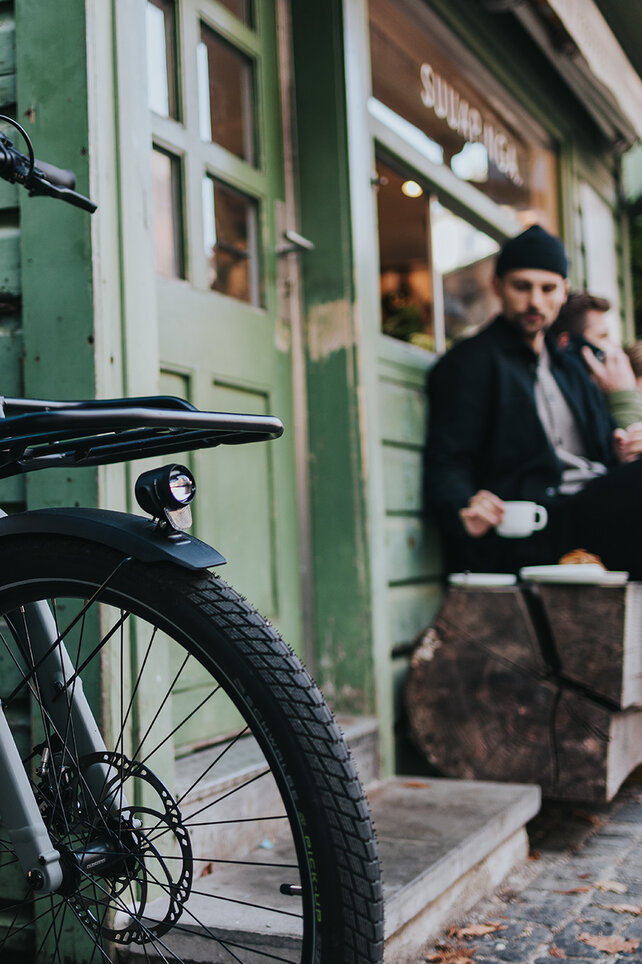 En modern, grå ellastcykel står i förgrunden framför ett café. I den suddiga bakgrunden sitter en man med keps på en träbänk och dricker ur en vit kopp.