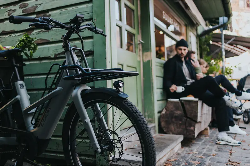 Ein modernes, graues eCargo Bike steht im Vordergrund vor einem Café. Im unscharfen Hintergrund sitzt ein Mann mit Mütze auf einer Holzbank und trinkt aus einer weißen Tasse.