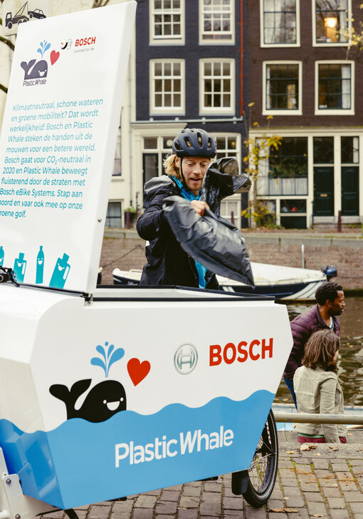 Een man staat voor een Cargo Bike met een laadbox. De laadbox is open en hij plaatst er een vuilniszak in. De box heeft een blauw-witte print met een zwarte walvis en het rode Bosch-logo.