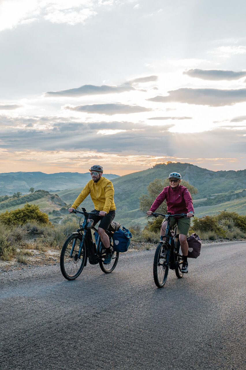 A woman and a man ride their eTrekking bikes along a road, with a wide landscape stretching out in the background.