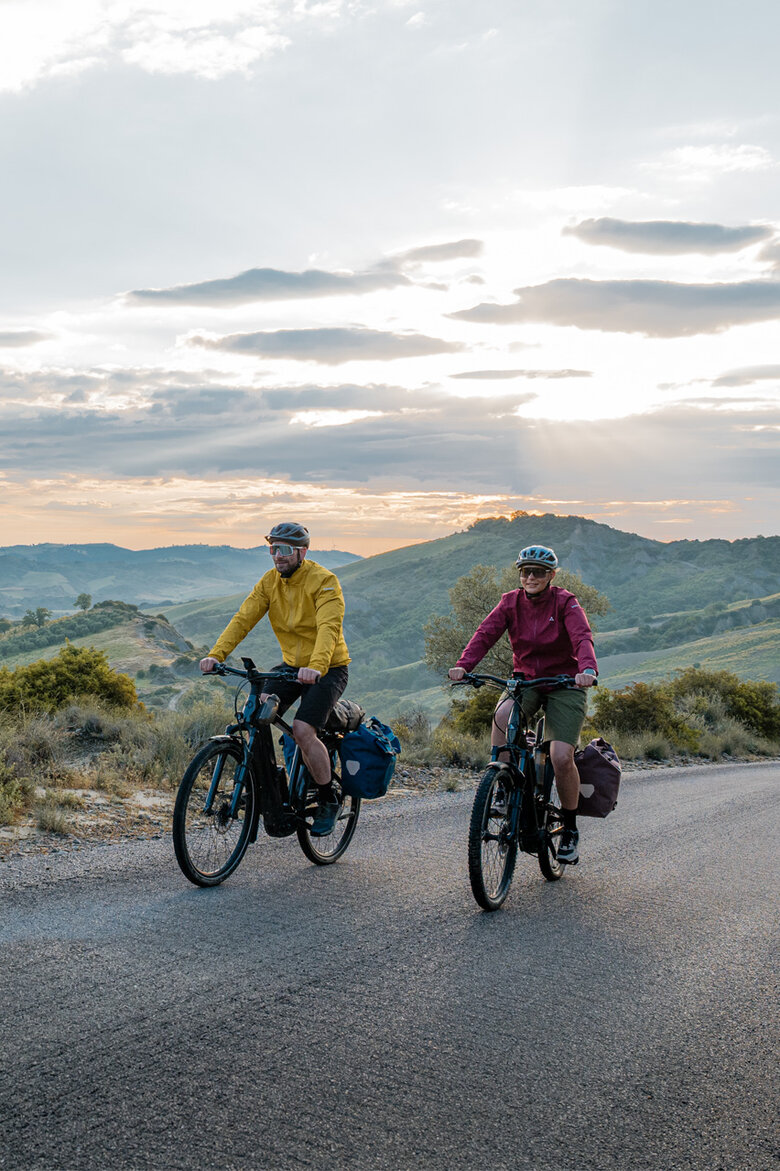 A woman and a man ride their eTrekking bikes along a road, with a wide landscape stretching out in the background.