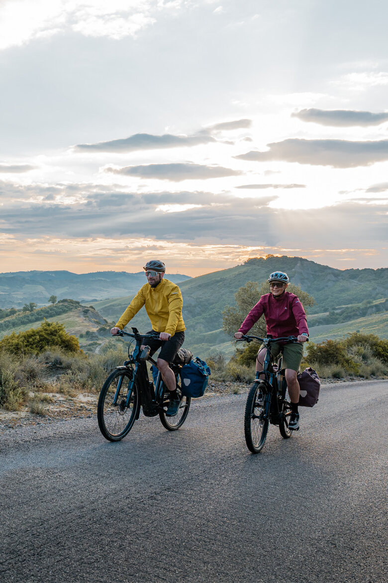 Eine Frau und ein Mann fahren mit ihren eTrekking-Bikes auf einer Straße, im Hintergrund ist eine weitläufige Landschaft zu sehen.