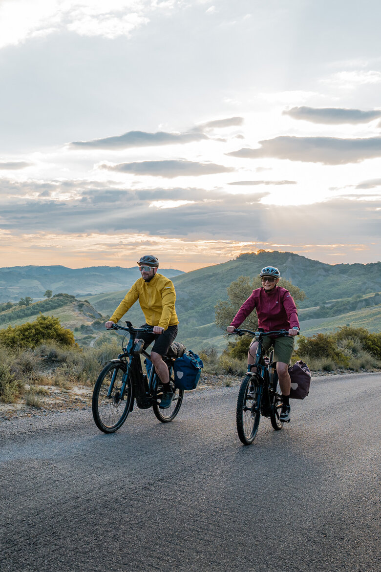 Eine Frau und ein Mann fahren mit ihren eTrekking-Bikes auf einer Straße, im Hintergrund ist eine weitläufige Landschaft zu sehen.