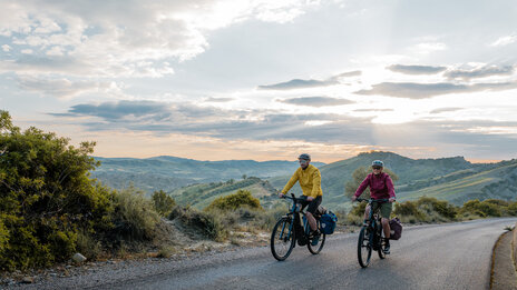 Eine Frau und ein Mann fahren mit ihren eTrekking-Bikes auf einer Straße, im Hintergrund ist eine weitläufige Landschaft zu sehen.
