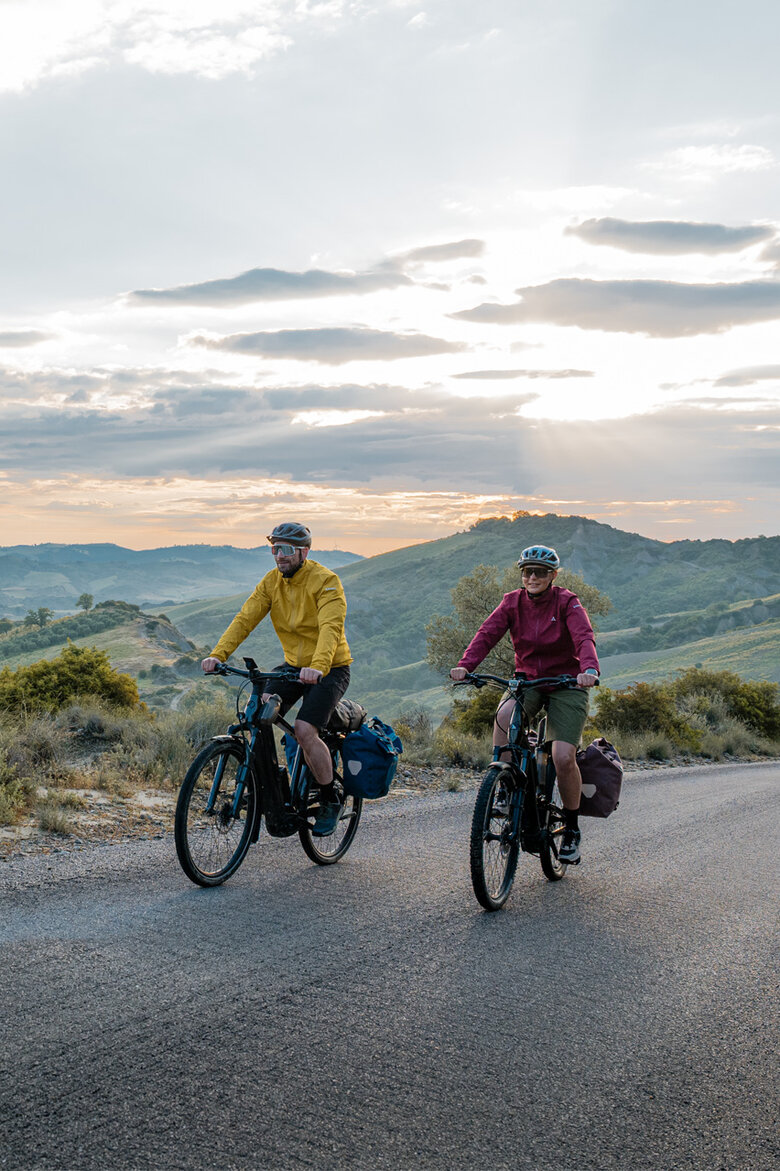 A woman and a man ride their eTrekking bikes along a road, with a wide landscape stretching out in the background.