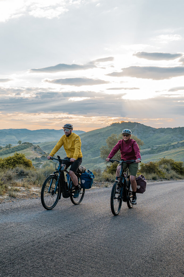Eine Frau und ein Mann fahren mit ihren eTrekking-Bikes auf einer Straße, im Hintergrund ist eine weitläufige Landschaft zu sehen.