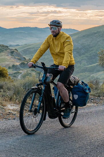 A woman and a man ride their eTrekking bikes along a road, with a wide landscape stretching out in the background.