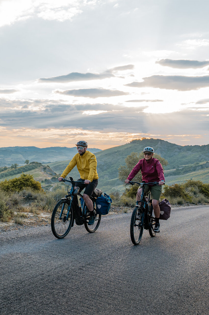 Eine Frau und ein Mann fahren mit ihren eTrekking-Bikes auf einer Straße, im Hintergrund ist eine weitläufige Landschaft zu sehen.
