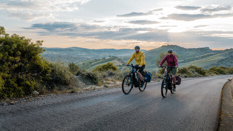 A woman and a man ride their eTrekking bikes along a road, with a wide landscape stretching out in the background.