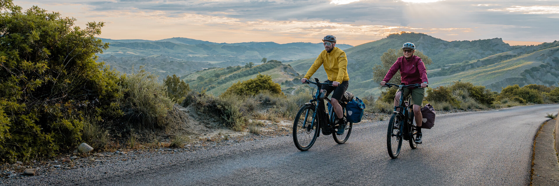 A woman and a man ride their eTrekking bikes along a road, with a wide landscape stretching out in the background.