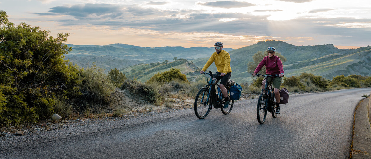 Eine Frau und ein Mann fahren mit ihren eTrekking-Bikes auf einer Straße, im Hintergrund ist eine weitläufige Landschaft zu sehen.