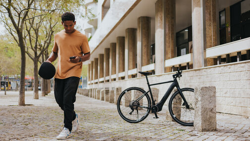 Un uomo si allontana da una eBike parcheggiata, controlla il suo smartphone e tiene un casco in mano.