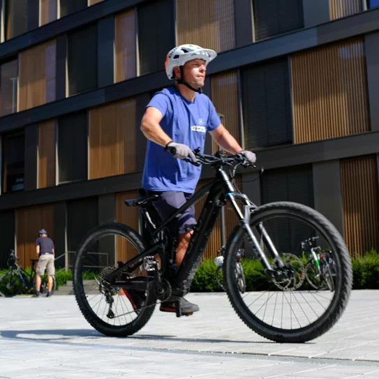 eBike Fahrer fährt auf dem Hof vor einem Gebäude, im Hintergrund holen zwei weitere Personen ihre eBikes.