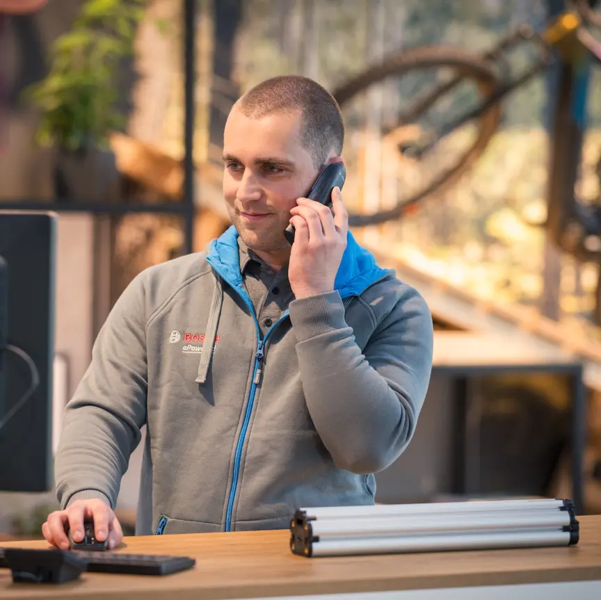 Eine Person steht an einem Schreibtisch und spricht am Telefon. Die Person trägt eine graue Bosch-Jacke und bedient gleichzeitig einen Computer. Im Hintergrund sind ein Fahrrad und Pflanzen zu sehen.
