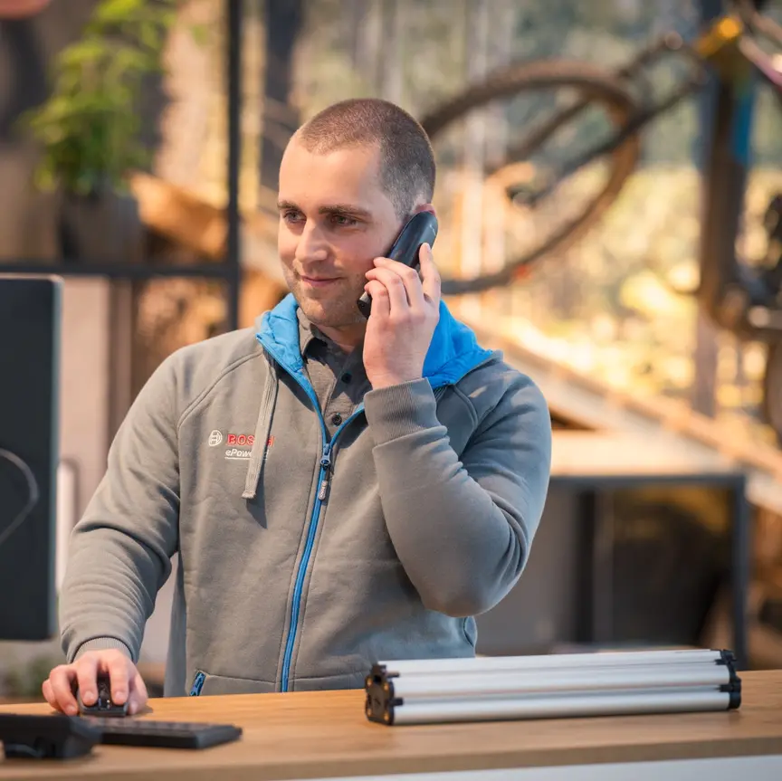 Eine Person steht an einem Schreibtisch und spricht am Telefon. Die Person trägt eine graue Bosch-Jacke und bedient gleichzeitig einen Computer. Im Hintergrund sind ein Fahrrad und Pflanzen zu sehen.