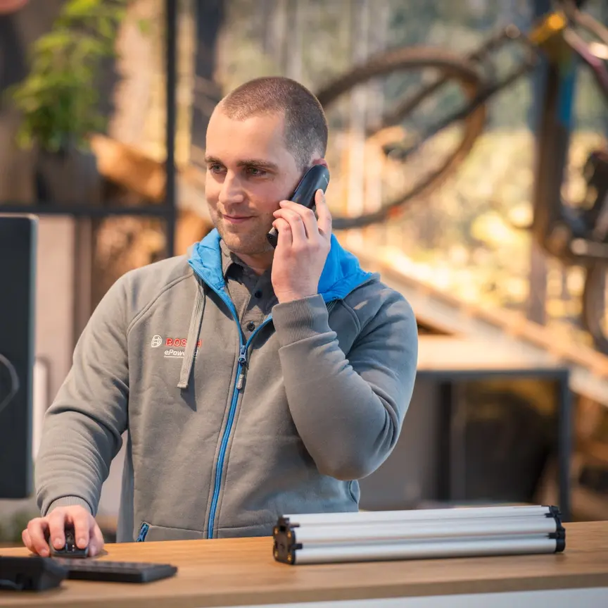 Eine Person steht an einem Schreibtisch und spricht am Telefon. Die Person trägt eine graue Bosch-Jacke und bedient gleichzeitig einen Computer. Im Hintergrund sind ein Fahrrad und Pflanzen zu sehen.