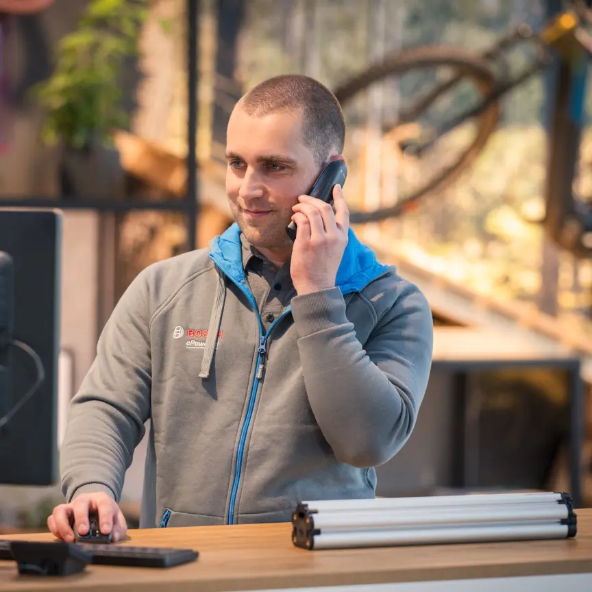 Une personne se tient à un bureau et parle au téléphone. Elle porte une veste Bosch grise et utilise simultanément un ordinateur. En arrière-plan, un vélo et des plantes sont visibles.