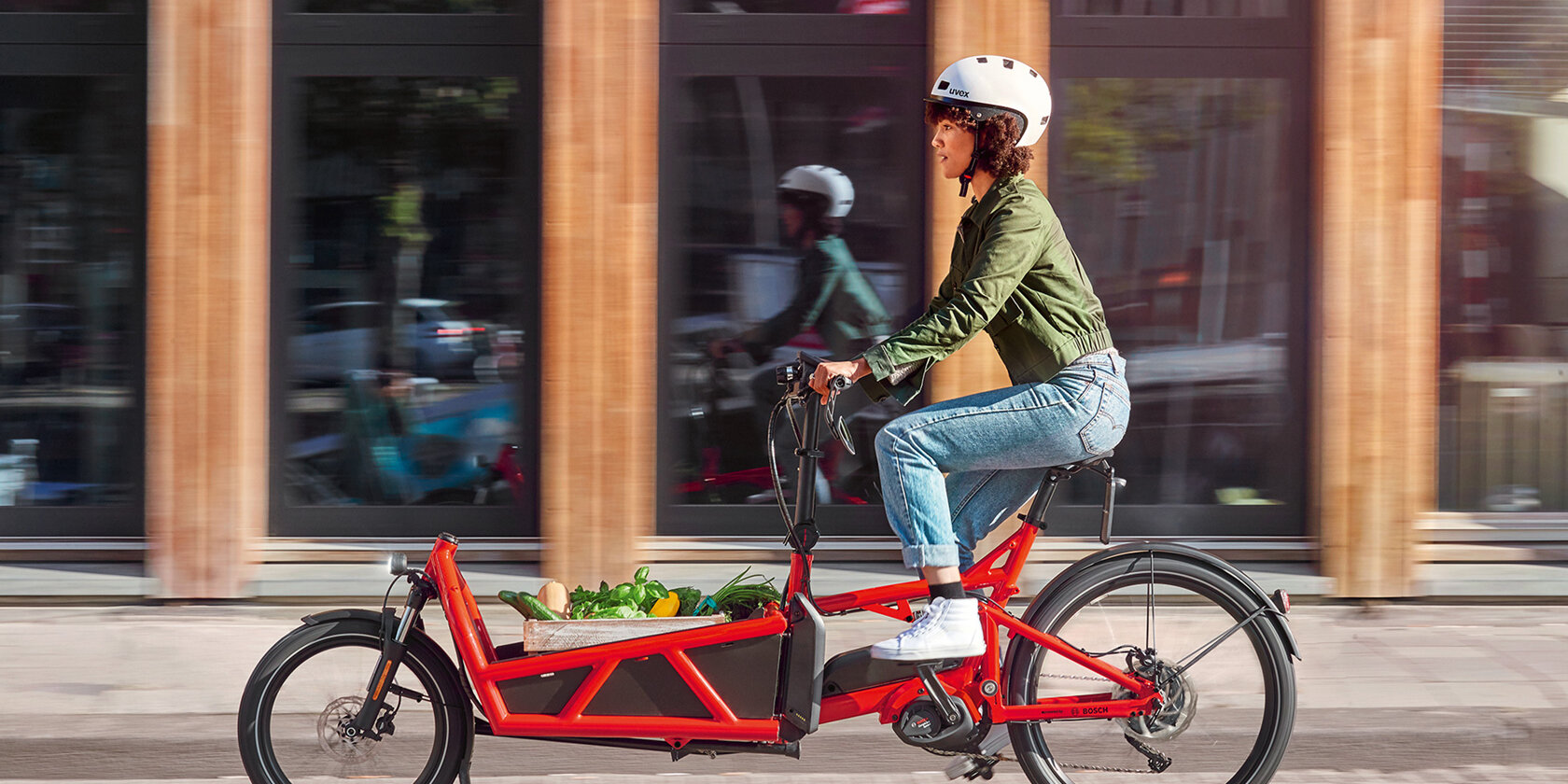 Frau mit eBike-Helm fährt auf rotem Lastenrad-Pedelec, das mit eineer Kiste Gemüse beladen ist, durch die Stadt