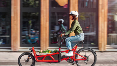 Frau mit eBike-Helm fährt auf rotem Lastenrad-Pedelec, das mit eineer Kiste Gemüse beladen ist, durch die Stadt
