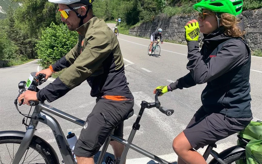 Un uomo e una donna su un tandem sono fermi ai margini di una strada, mentre altri due conducenti di eBike passano lungo la stessa via.