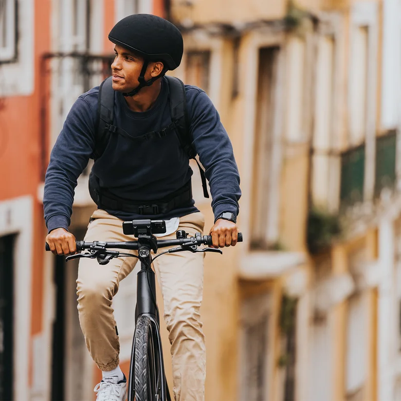 A man rides his eBike through an alley, glancing to the side. A smartphone is mounted on the eBike, with houses visible in the background.