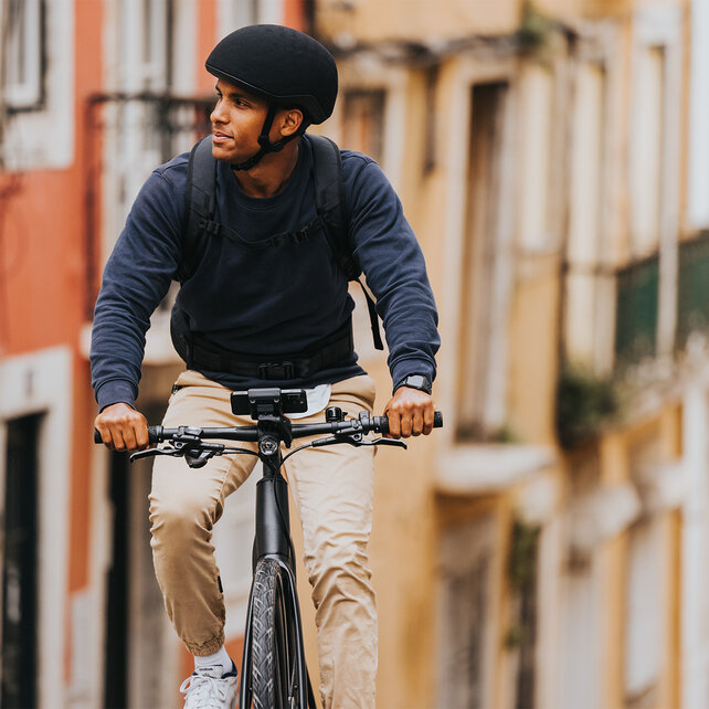A man rides his eBike through an alley, glancing to the side. A smartphone is mounted on the eBike, with houses visible in the background.
