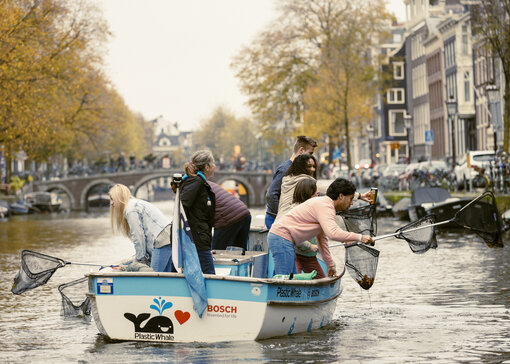 Meerdere mensen zitten op een bootje op een gracht en vissen met schepnetten dingen uit het water.