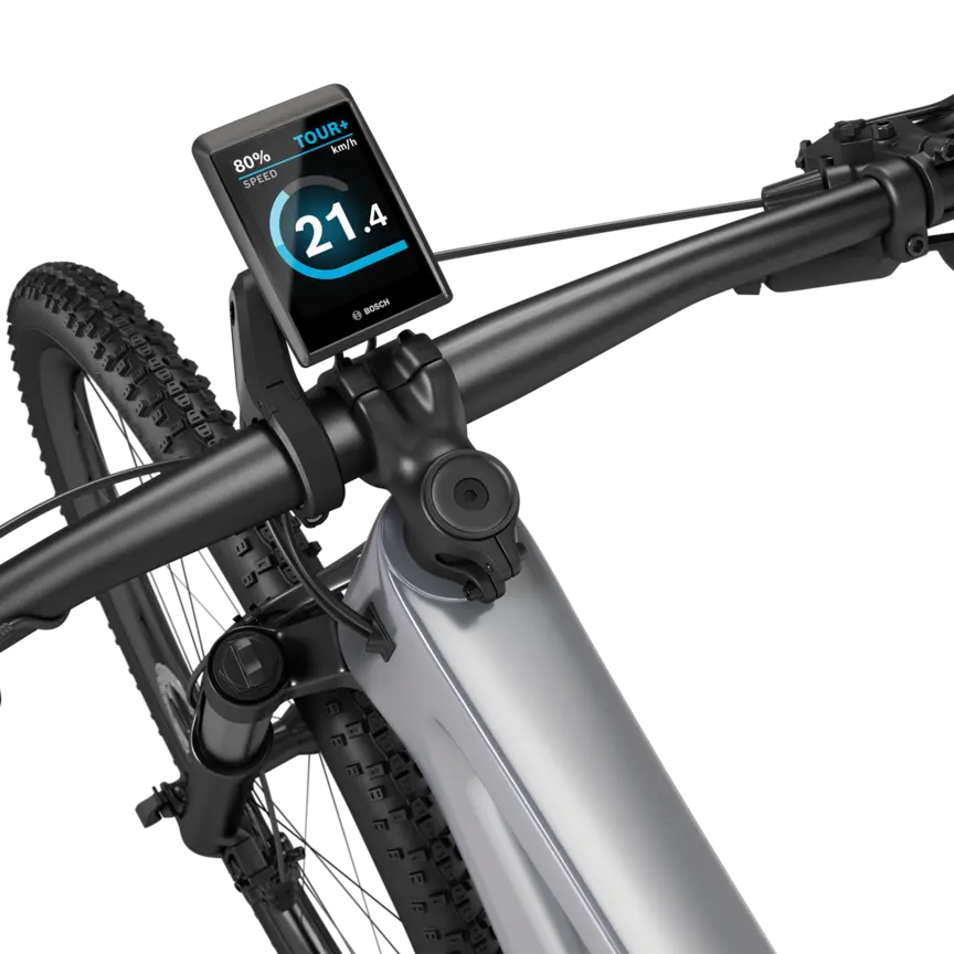 A silver eMountain bike with a black handlebar stands isolated against a white background. A Kiox 500 is mounted on the handlebar. The display shows the speed in km/h, the battery level, and the riding mode.