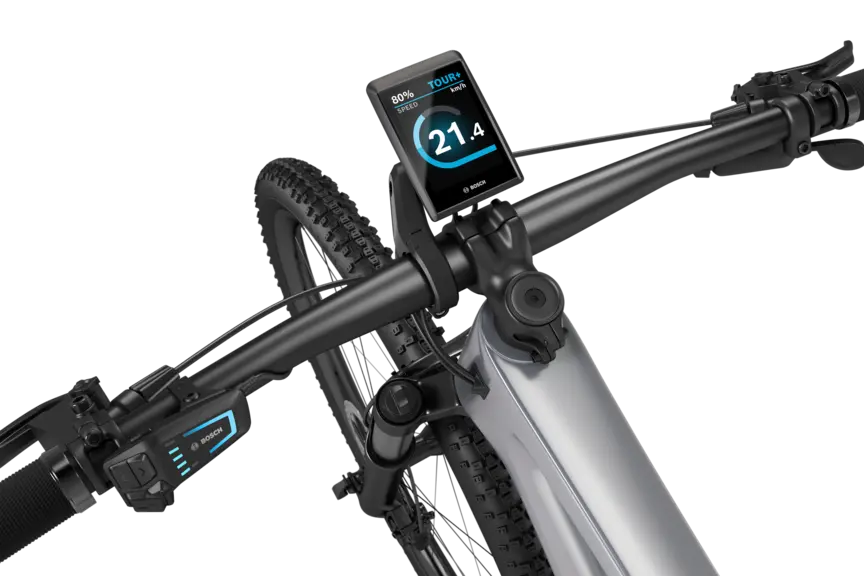 A silver eMountain bike with a black handlebar stands isolated against a white background. A Kiox 500 is mounted on the handlebar. The display shows the speed in km/h, the battery level, and the riding mode.