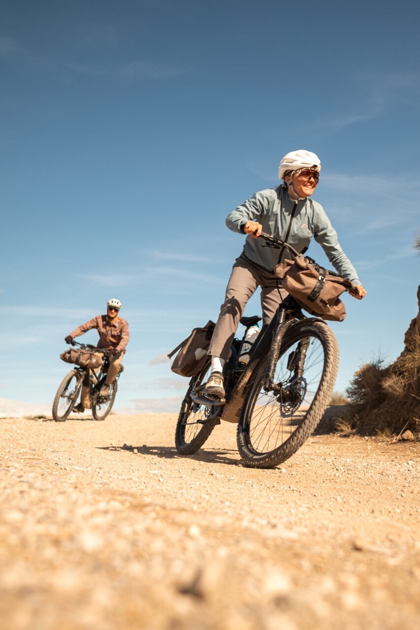 Zwei Personen auf bepackten eBikes auf einem Schotterweg. Die Frau im Vordergrund lächelt, während sie in einer trockenen, hügeligen Landschaft fährt.