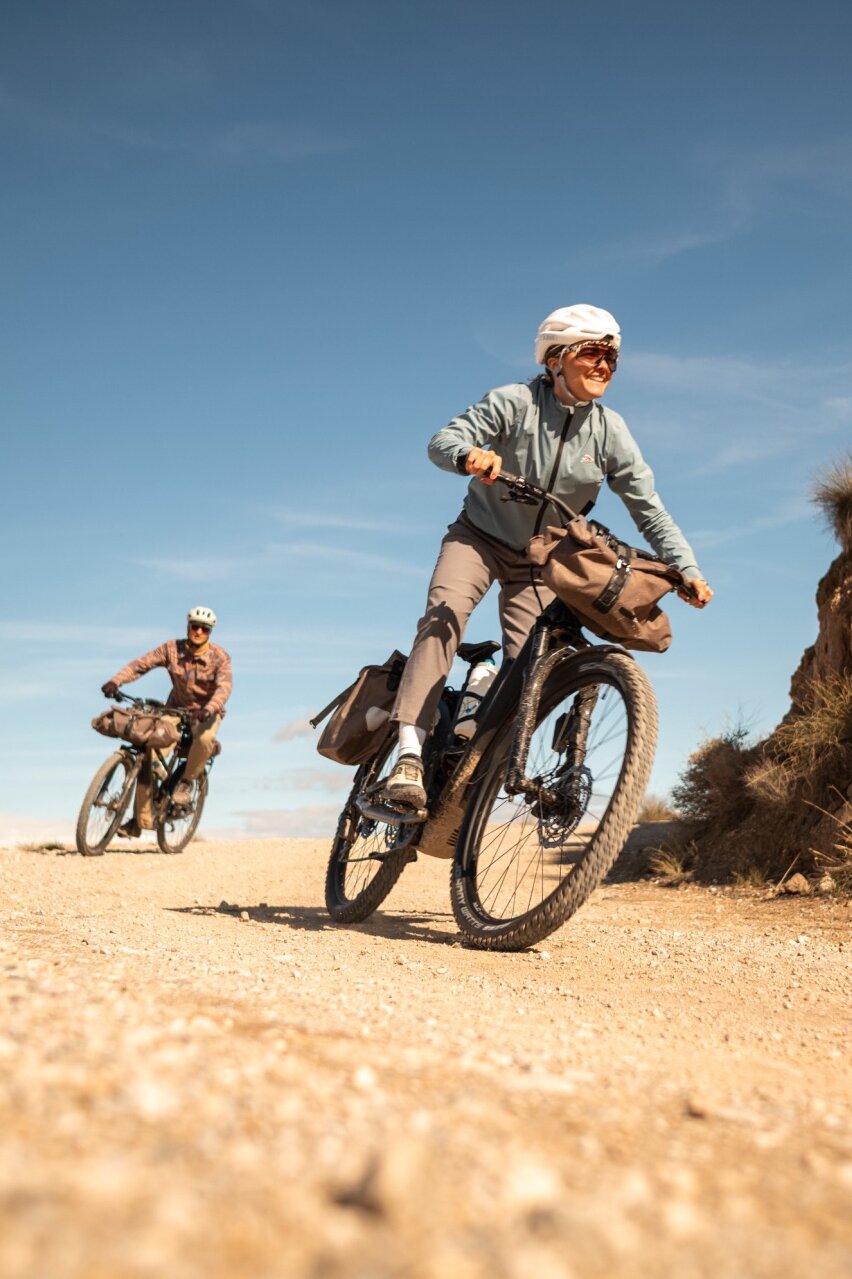 Zwei Personen auf bepackten eBikes auf einem Schotterweg. Die Frau im Vordergrund lächelt, während sie in einer trockenen, hügeligen Landschaft fährt.
