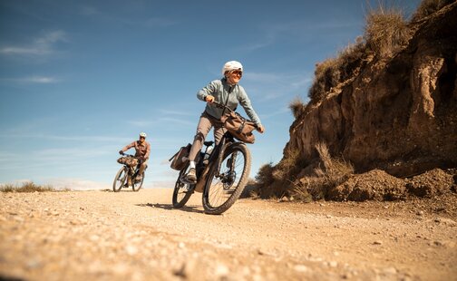 Två personer på utrustade eBikes på en grusväg. Kvinnan i förgrunden ler medan hon cyklar genom ett torrt, kuperat landskap.