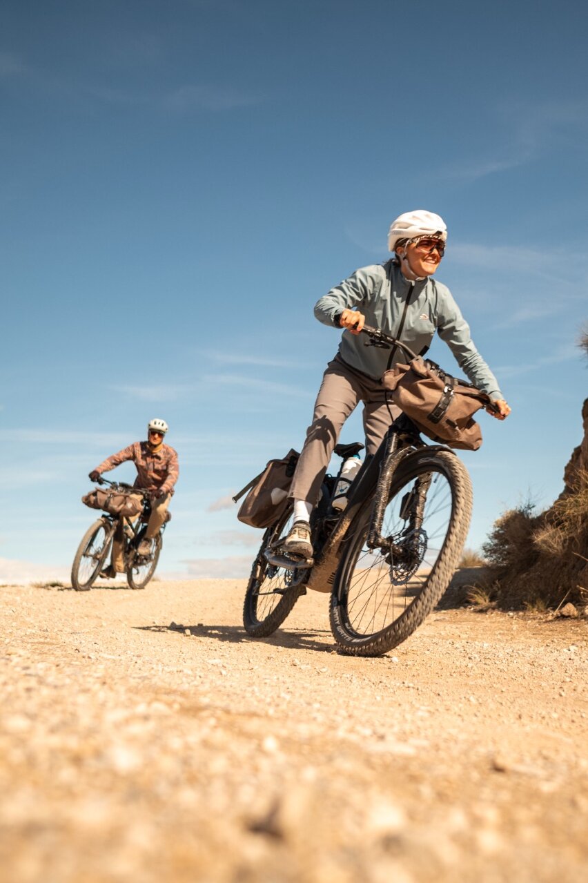 Two people on loaded eBikes ride along a gravel path. The woman in the foreground smiles as she rides through a dry, hilly landscape.