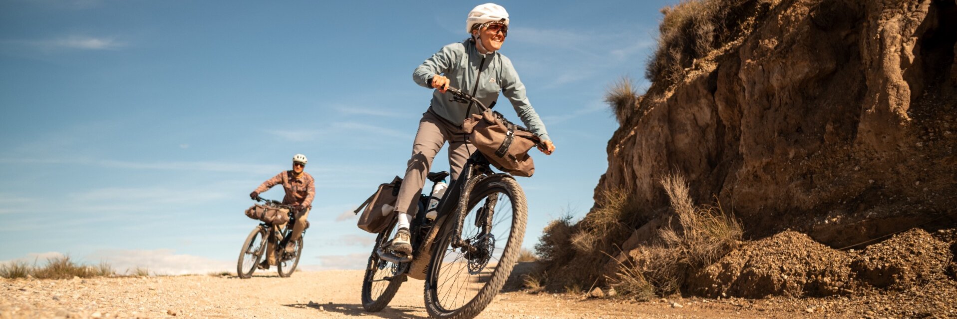 Dos personas sobre eBikes cargadas recorren un camino de grava. La mujer en primer plano sonríe mientras pedalea por un paisaje seco y montañoso.