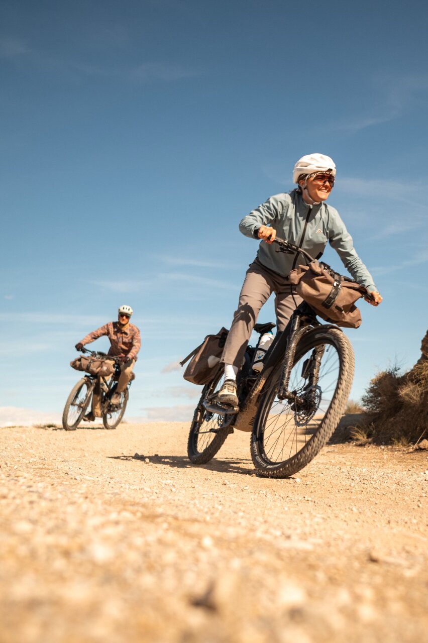 Zwei Personen auf bepackten eBikes auf einem Schotterweg. Die Frau im Vordergrund lächelt, während sie in einer trockenen, hügeligen Landschaft fährt.