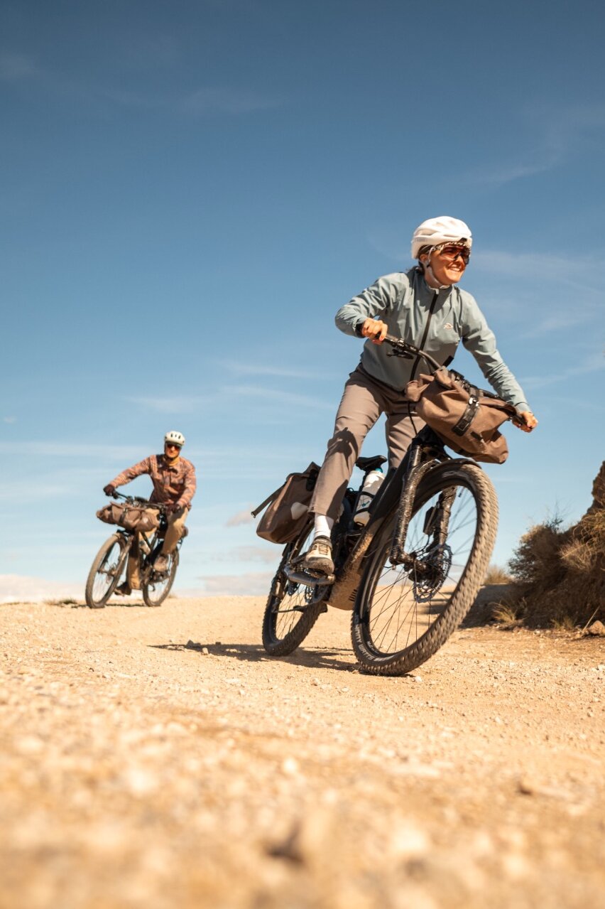 Zwei Personen auf bepackten eBikes auf einem Schotterweg. Die Frau im Vordergrund lächelt, während sie in einer trockenen, hügeligen Landschaft fährt.