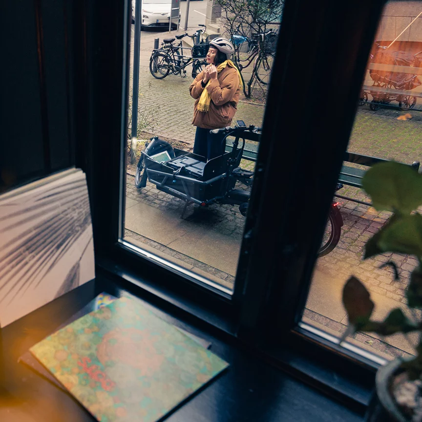 Jezdkyně na nákladní elektrokolo si připíná cyklistickou helmu, snímek je pořízený přes skleněnou výlohu kavárny.