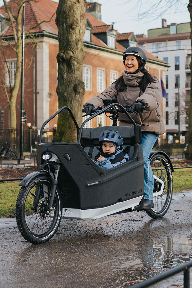 Eine Person fährt ein Lasten-E-Bike auf einem nassen Weg in einer städtischen Umgebung. Im Lastenrad sitzt ein Kind, das einen blauen Helm trägt. Im Hintergrund sind Gebäude, Bäume und Flaggen zu sehen.