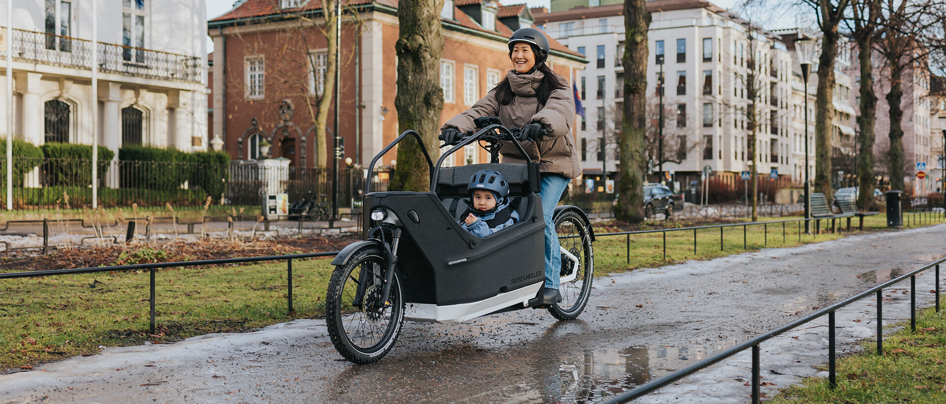 Eine Person fährt ein Lasten-E-Bike auf einem nassen Weg in einer städtischen Umgebung. Im Lastenrad sitzt ein Kind, das einen blauen Helm trägt. Im Hintergrund sind Gebäude, Bäume und Flaggen zu sehen.