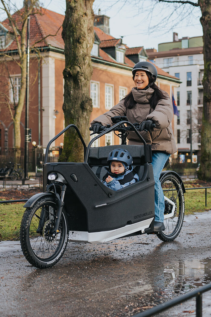 Eine Person fährt ein Lasten-E-Bike auf einem nassen Weg in einer städtischen Umgebung. Im Lastenrad sitzt ein Kind, das einen blauen Helm trägt. Im Hintergrund sind Gebäude, Bäume und Flaggen zu sehen.