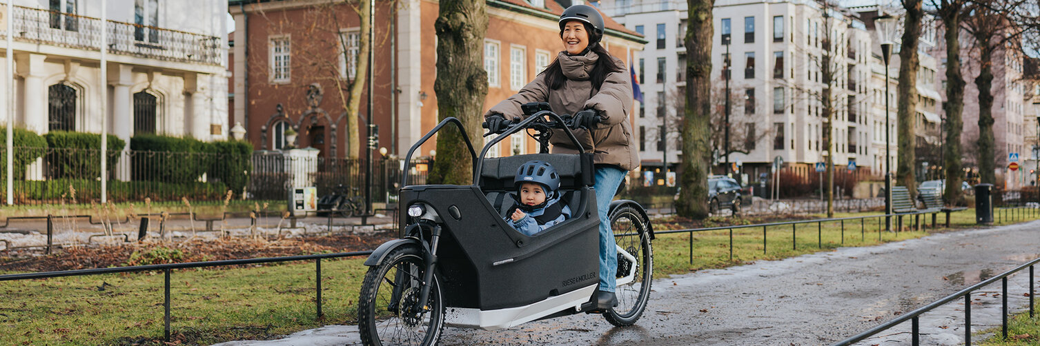 A person rides a cargo e-bike on a wet path in an urban environment. In the cargo bikes, a child wearing a blue helmet is seated. In the background, you can see buildings, trees, and flags.
