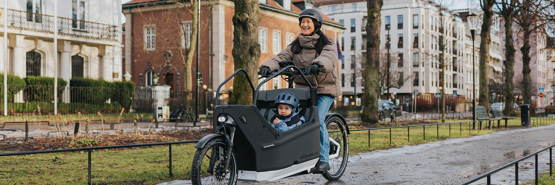Een persoon rijdt op een vracht-e-bike over een nat pad in een stedelijke omgeving. In de bakfiets zit een kind met een blauwe helm. Op de achtergrond zie je gebouwen, bomen en vlaggen.