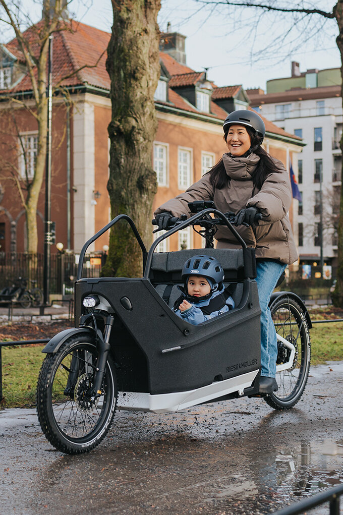 Eine Person fährt ein Lasten-E-Bike auf einem nassen Weg in einer städtischen Umgebung. Im Lastenrad sitzt ein Kind, das einen blauen Helm trägt. Im Hintergrund sind Gebäude, Bäume und Flaggen zu sehen.