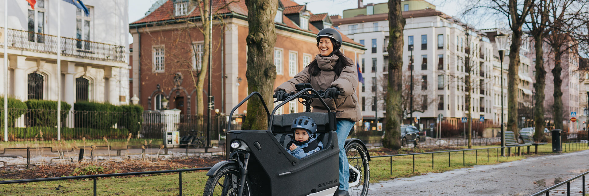 A person rides a cargo e-bike on a wet path in an urban environment. In the cargo bikes, a child wearing a blue helmet is seated. In the background, you can see buildings, trees, and flags.