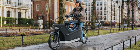 Eine Person fährt ein Lasten-E-Bike auf einem nassen Weg in einer städtischen Umgebung. Im Lastenrad sitzt ein Kind, das einen blauen Helm trägt. Im Hintergrund sind Gebäude, Bäume und Flaggen zu sehen.