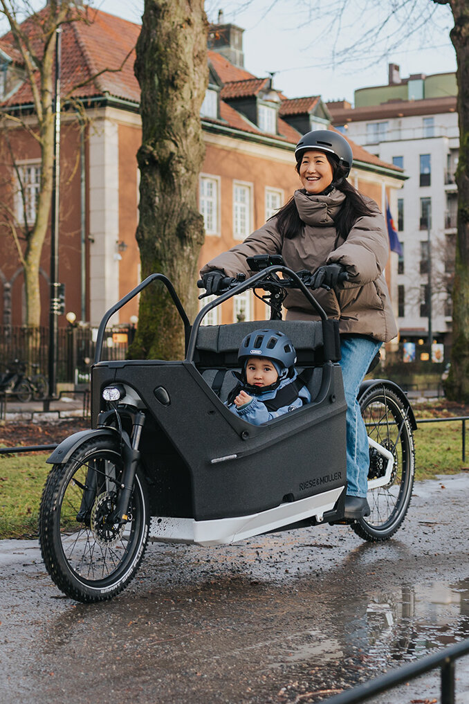 Eine Person fährt ein Lasten-E-Bike auf einem nassen Weg in einer städtischen Umgebung. Im Lastenrad sitzt ein Kind, das einen blauen Helm trägt. Im Hintergrund sind Gebäude, Bäume und Flaggen zu sehen.