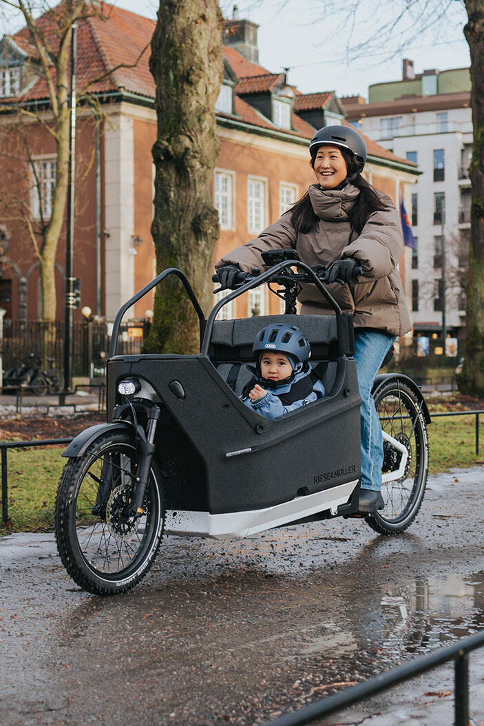 Eine Person fährt ein Lasten-E-Bike auf einem nassen Weg in einer städtischen Umgebung. Im Lastenrad sitzt ein Kind, das einen blauen Helm trägt. Im Hintergrund sind Gebäude, Bäume und Flaggen zu sehen.
