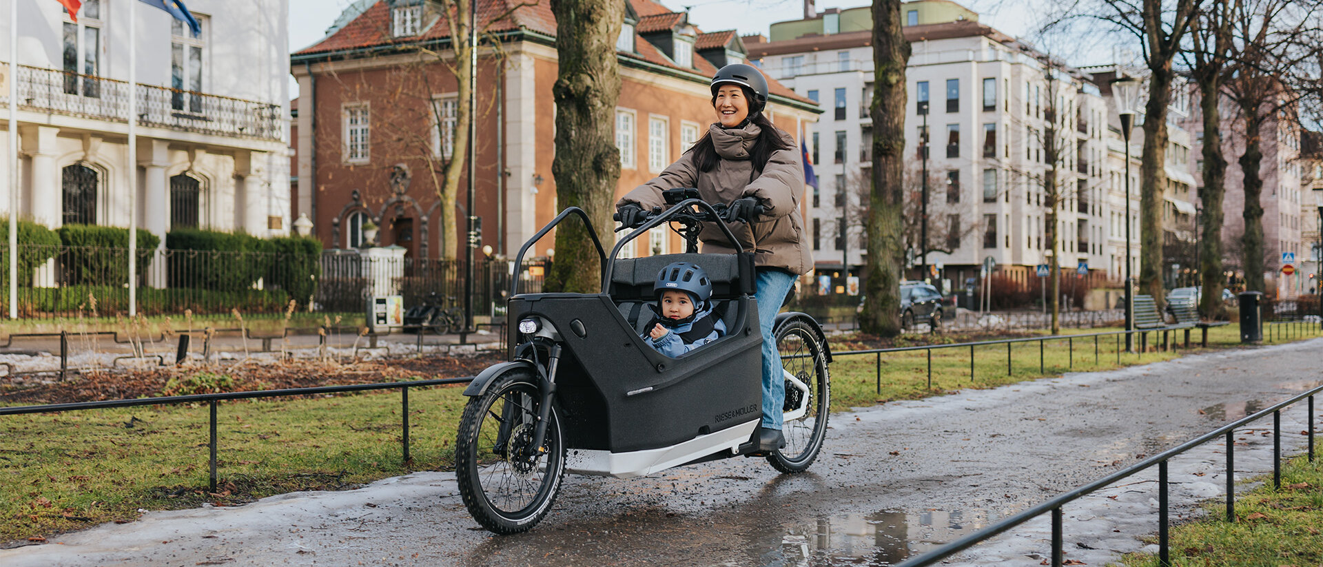 Eine Person fährt ein Lasten-E-Bike auf einem nassen Weg in einer städtischen Umgebung. Im Lastenrad sitzt ein Kind, das einen blauen Helm trägt. Im Hintergrund sind Gebäude, Bäume und Flaggen zu sehen.