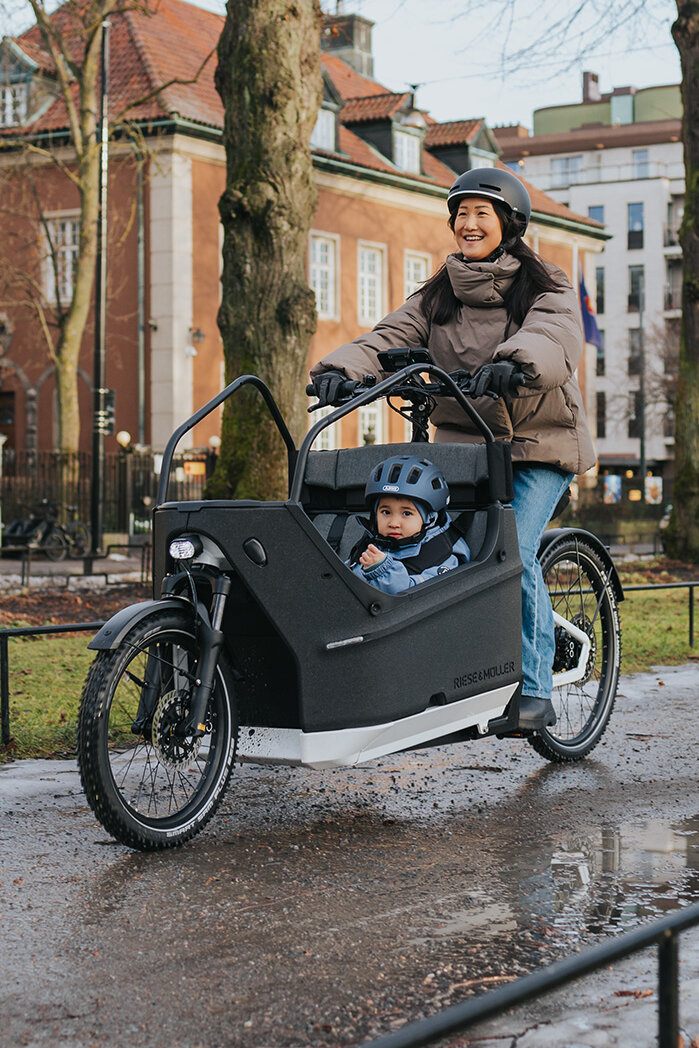 A person rides a cargo e-bike on a wet path in an urban environment. In the cargo bikes, a child wearing a blue helmet is seated. In the background, you can see buildings, trees, and flags.