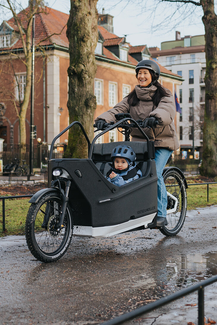 Eine Person fährt ein Lasten-E-Bike auf einem nassen Weg in einer städtischen Umgebung. Im Lastenrad sitzt ein Kind, das einen blauen Helm trägt. Im Hintergrund sind Gebäude, Bäume und Flaggen zu sehen.
